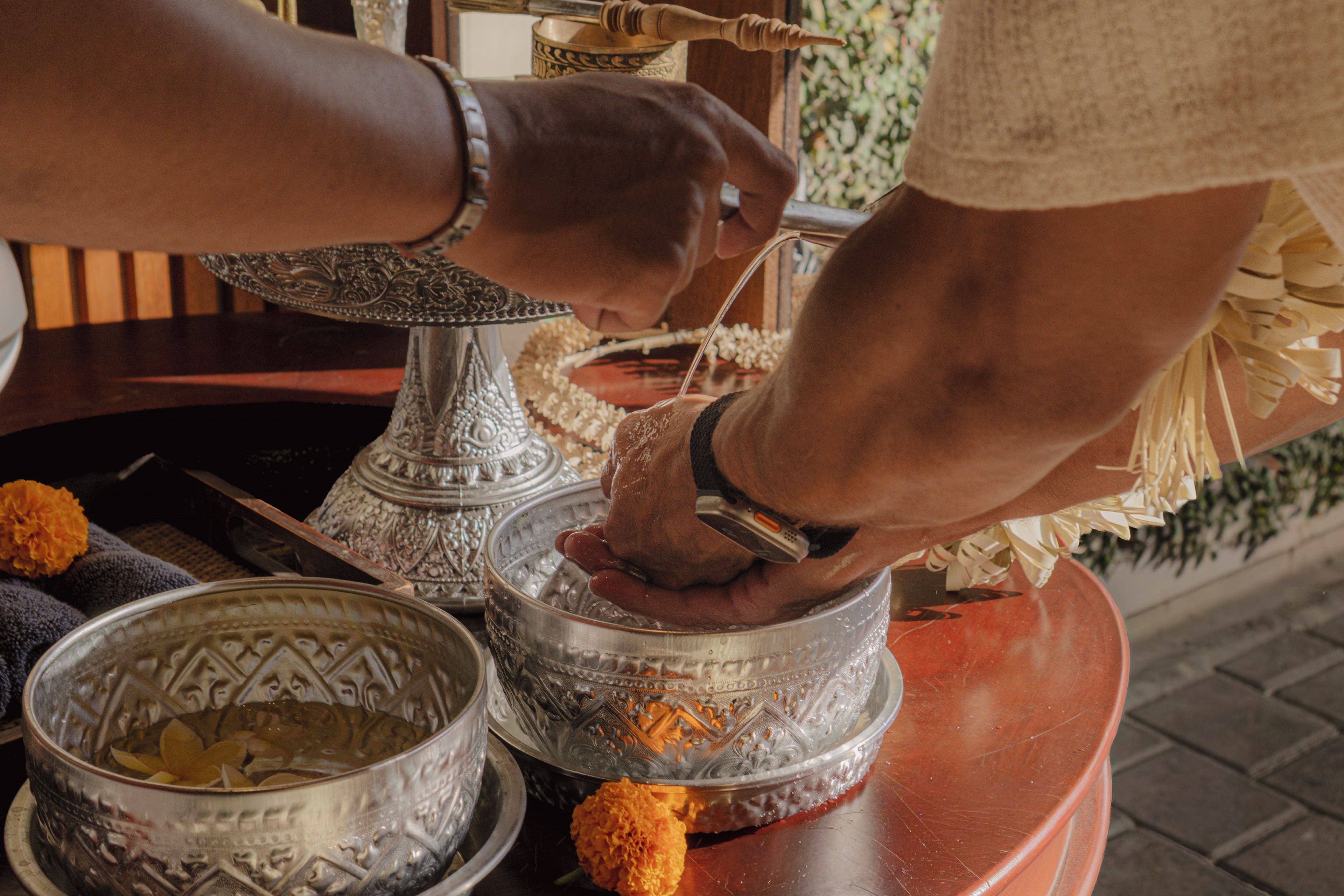 A person performing a ritual with a silver bowl and a brass container, surrounded by ornate silver vessels and marigold flowers.
