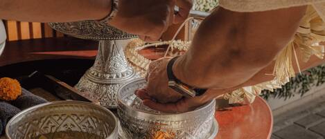 A person performing a ritual with a silver bowl and a brass container, surrounded by ornate silver vessels and marigold flowers.