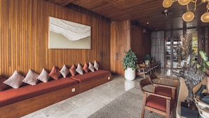 A modern interior with a wooden panel wall, a red sofa with patterned cushions, a large potted plant, and a dining area with wooden chairs and a table. - Sana Vie Villa Seminyak by Ini Vie Hospitality (Seminyak)