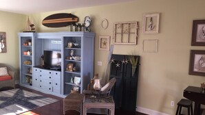 A living room with a blue entertainment center, a wicker chair, a glass coffee table, and a wooden table with a stool. - Luxury Condo - Vofp - "Featured in Delaware Today" (Lewes)