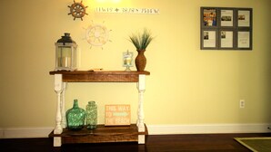 A wooden console table with a sign reading "THIS WAY TO THE BEACH," a ship's wheel, a lantern, and a framed bulletin board on a yellow wall. - Luxury Condo - Vofp - "Featured in Delaware Today" (Lewes)
