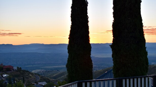 Vista de un paisaje con dos árboles altos en primer plano, una barandilla y una lejana vista de un valle y montañas.