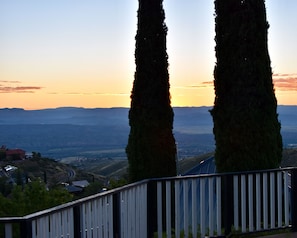 Une vue d’un paysage avec deux grands arbres au premier plan, une balustrade, et une vue lointaine d’une vallée et de montagnes.