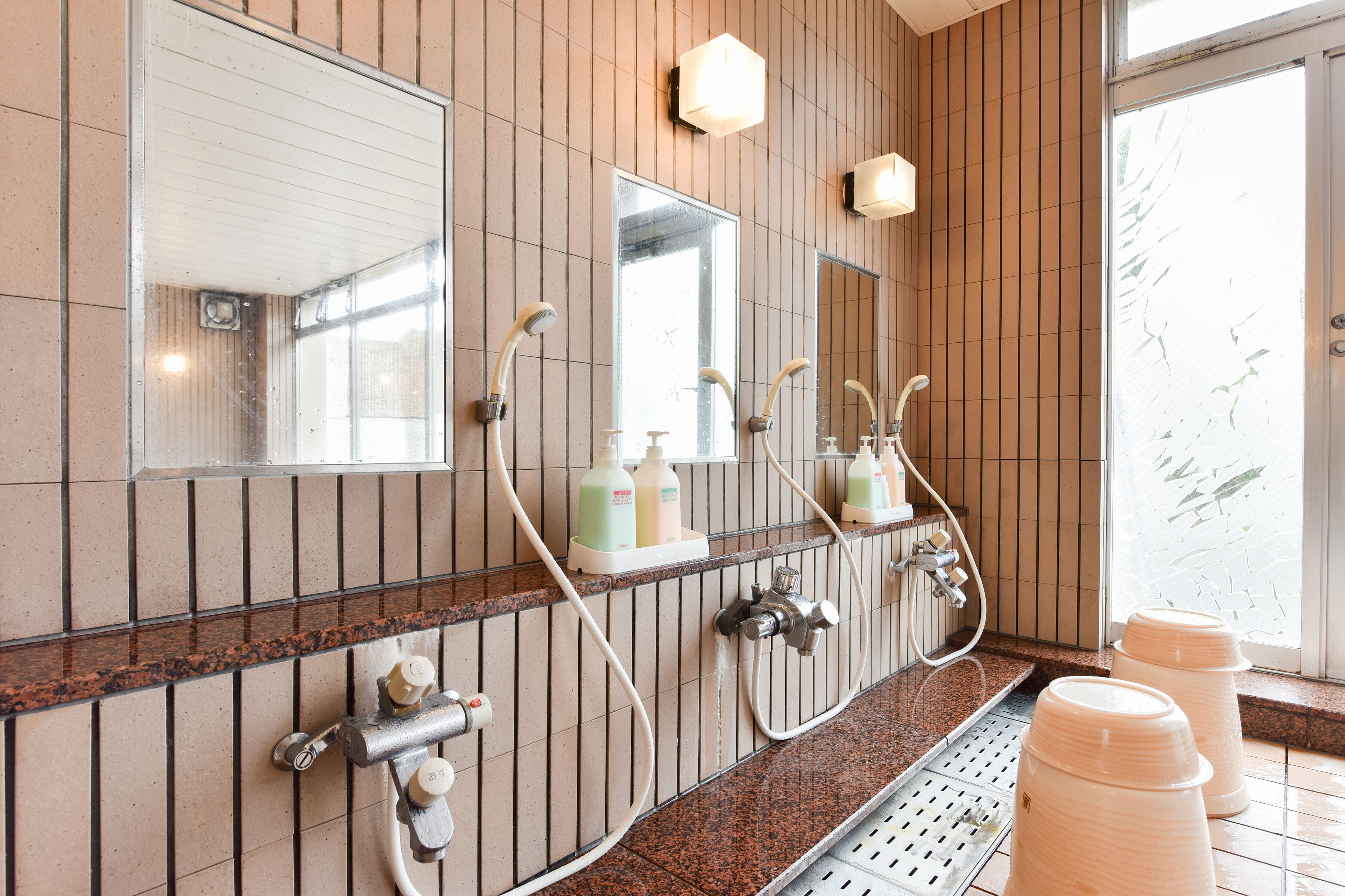 A bathroom with a long sink, multiple faucets, and mirrors. There are bottles of soap and a showerhead visible.