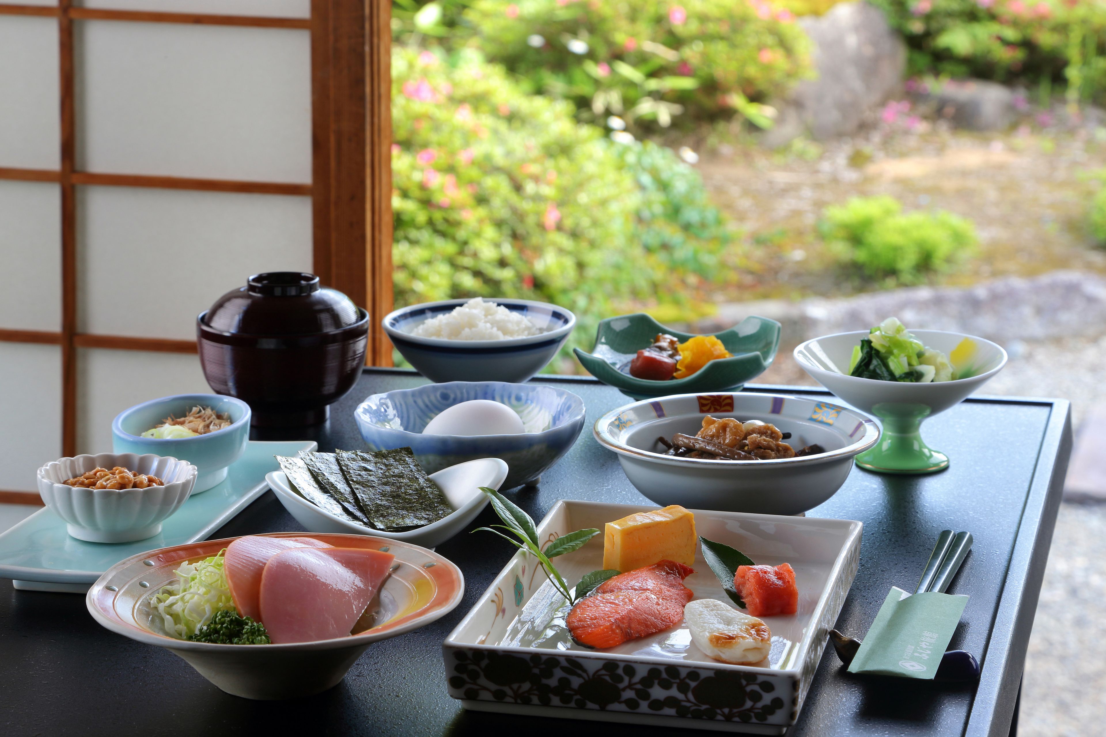 Un montaje tradicional de comida japonesa con varios platos sobre una mesa, incluyendo arroz, pescado y verduras, con vistas a un jardín al fondo.