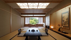 A traditional Japanese room with tatami flooring, a low table with cushions, and a view of a forested area through large windows. - Bekkan Fujiya Ryokan (Kaminoyama)