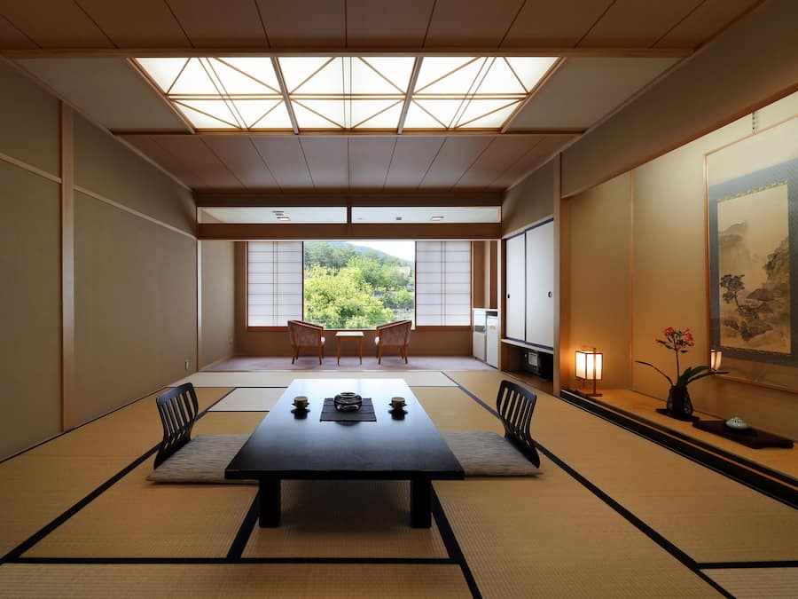 A traditional Japanese room with tatami flooring, a low table with cushions, and a view of a forested area through large windows.
