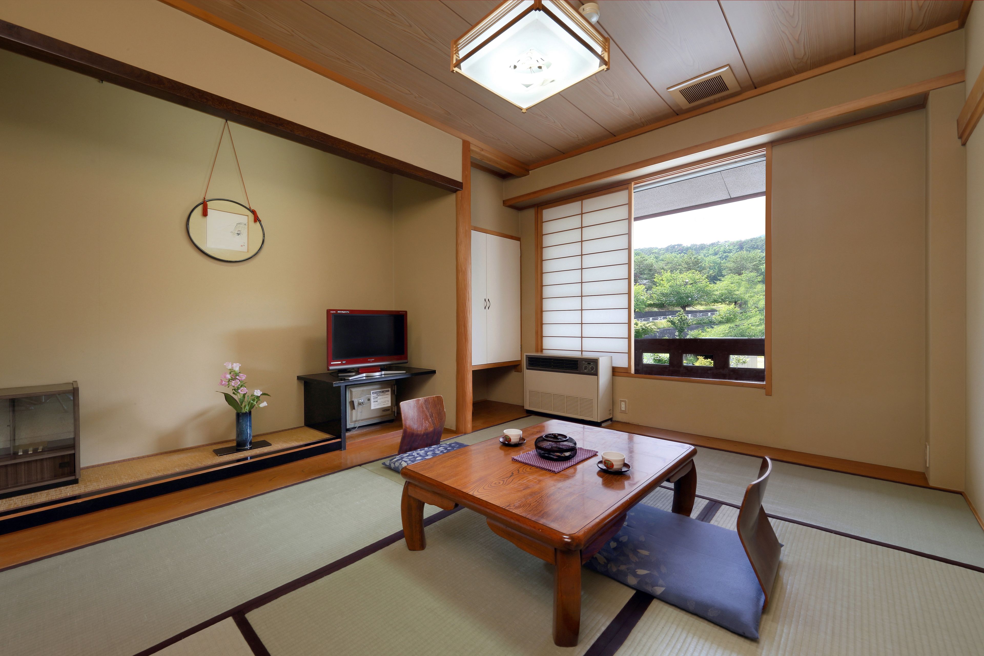 A traditional Japanese room with tatami flooring, a low wooden table, a television, and a view of greenery outside.