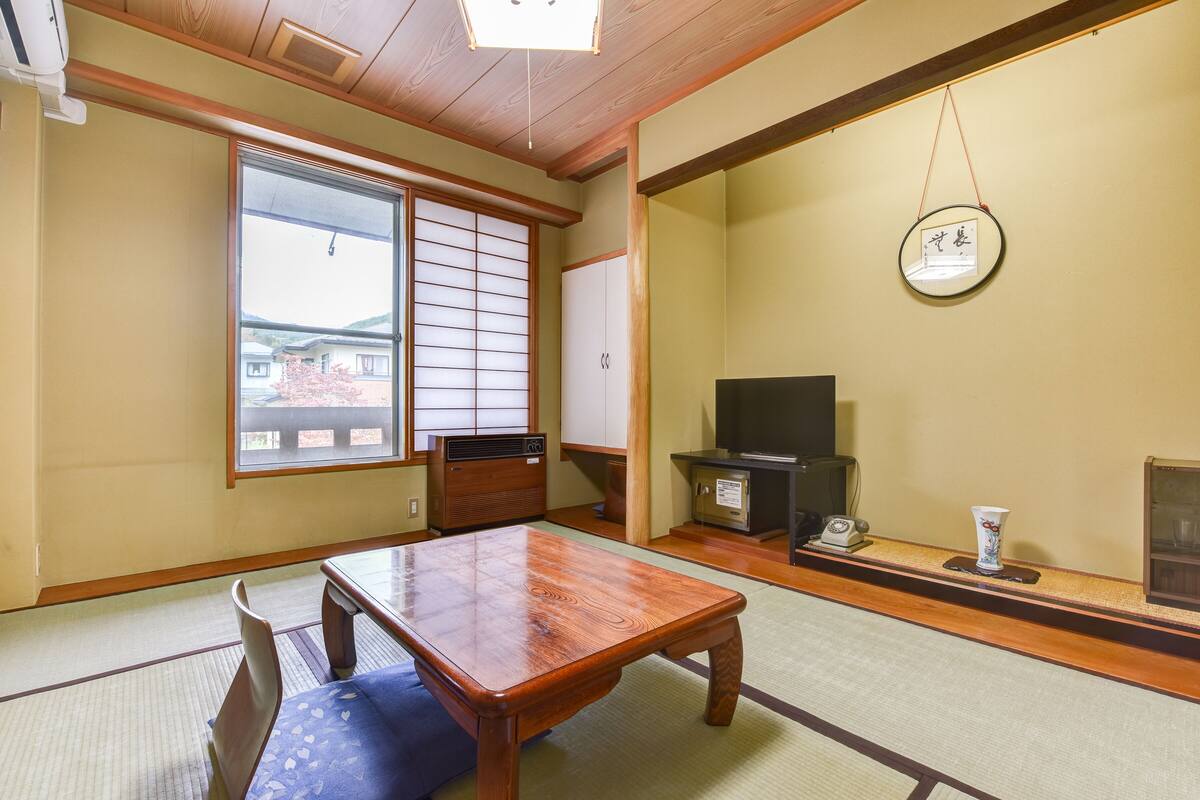 Ein traditioneller japanischer Raum mit Tatami-Bodenbelag, einem Holztisch, einem Fernseher und einer Schiebetür.