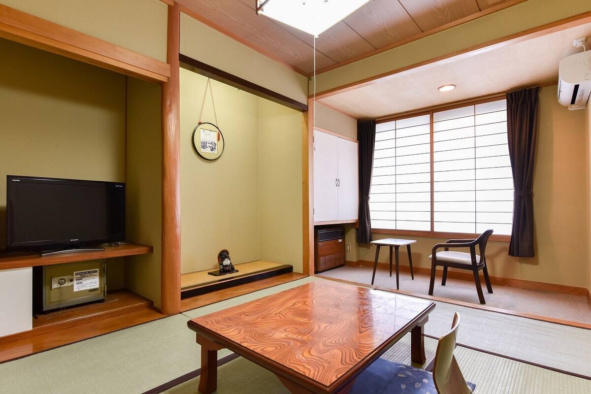 Ein traditioneller japanischer Raum mit Tatami-Boden, einem Fernseher, einem kleinen Schrein, einer Schiebetür und Blick ins Freie.