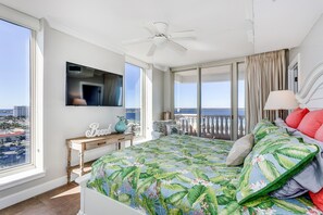 Un dormitorio con una cama grande, un ventilador de techo, un televisor montado en la pared y vistas de la ciudad a través de amplias ventanas.