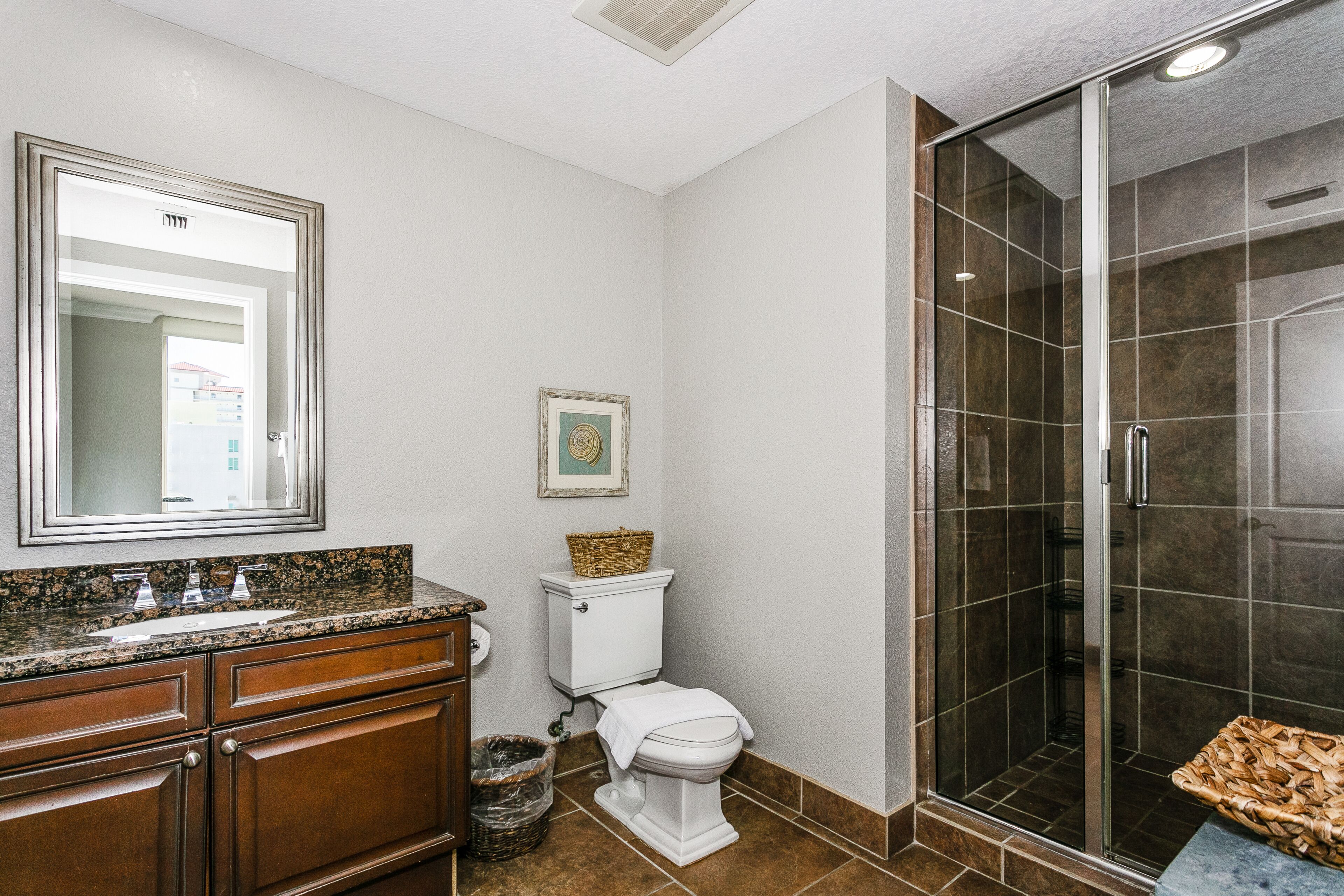Une salle de bain avec des toilettes blanches, un grand miroir, un comptoir en granit, des meubles en bois et un espace douche doté de portes en verre.