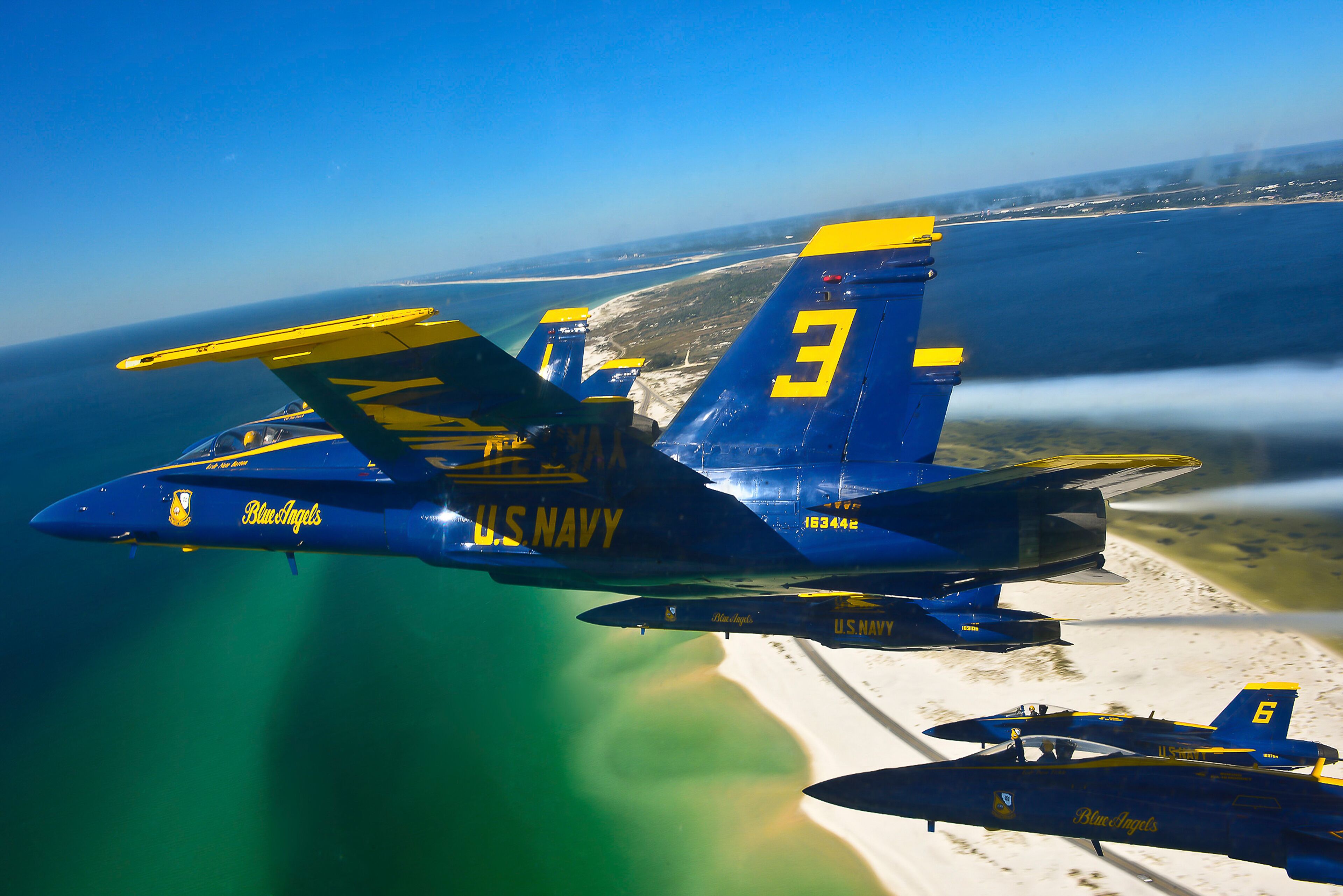 Un avión de los Blue Angels de la Marina estadounidense, con el número 3, volando sobre una playa con cielos despejados.
