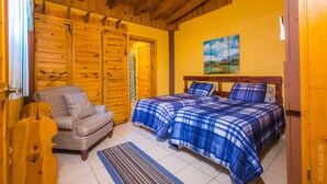 Een kamer met houten wanden en plafond, een bed met blauw beddengoed, een stoel en een schilderij aan de muur.