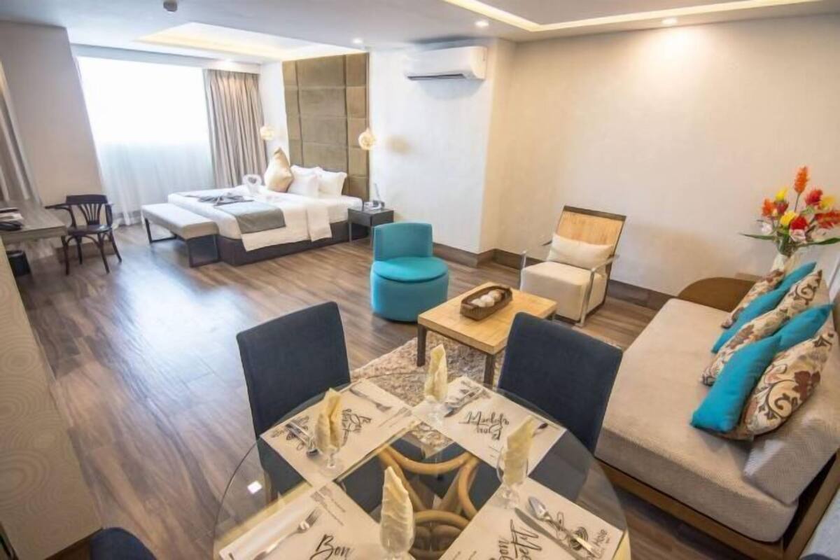 A modern hotel room with a sofa, a dining table set with a white tablecloth, and a small side table with a vase of flowers.