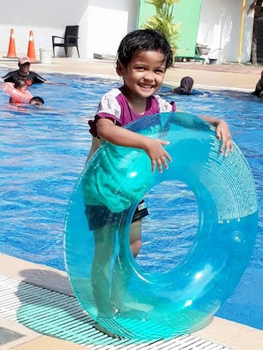 Un enfant tenant un grand anneau gonflable dans une zone de piscine, avec un sol carrelé et quelques cônes de signalisation orange en arrière-plan.