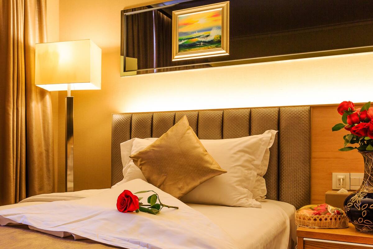 A hotel room with a neatly made bed, a bedside lamp, a framed picture above the bed, and a vase with flowers on the nightstand.
