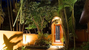 A well-lit entrance with a gated door, surrounded by lush tropical plants and a pathway leading to a vehicle. - Sazana Villa Resort (Koh Samui)