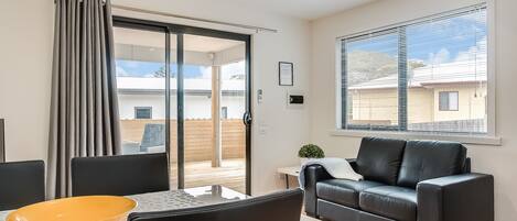 A modern living room with a glass door leading to a balcony, a black leather sofa, a dining table with chairs, and a large window with blinds.