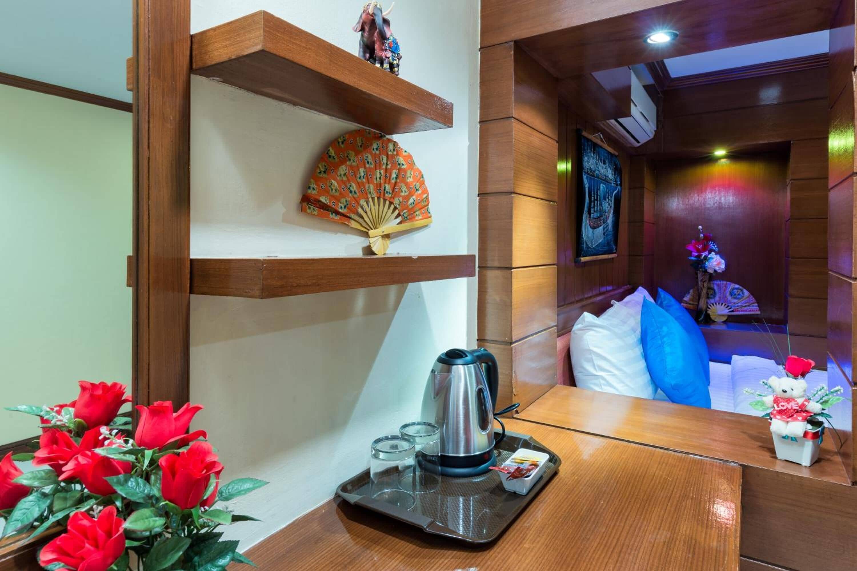 A hotel room with a wooden table, a kettle, a tray with glasses, a decorative fan, and a vase with red flowers.
