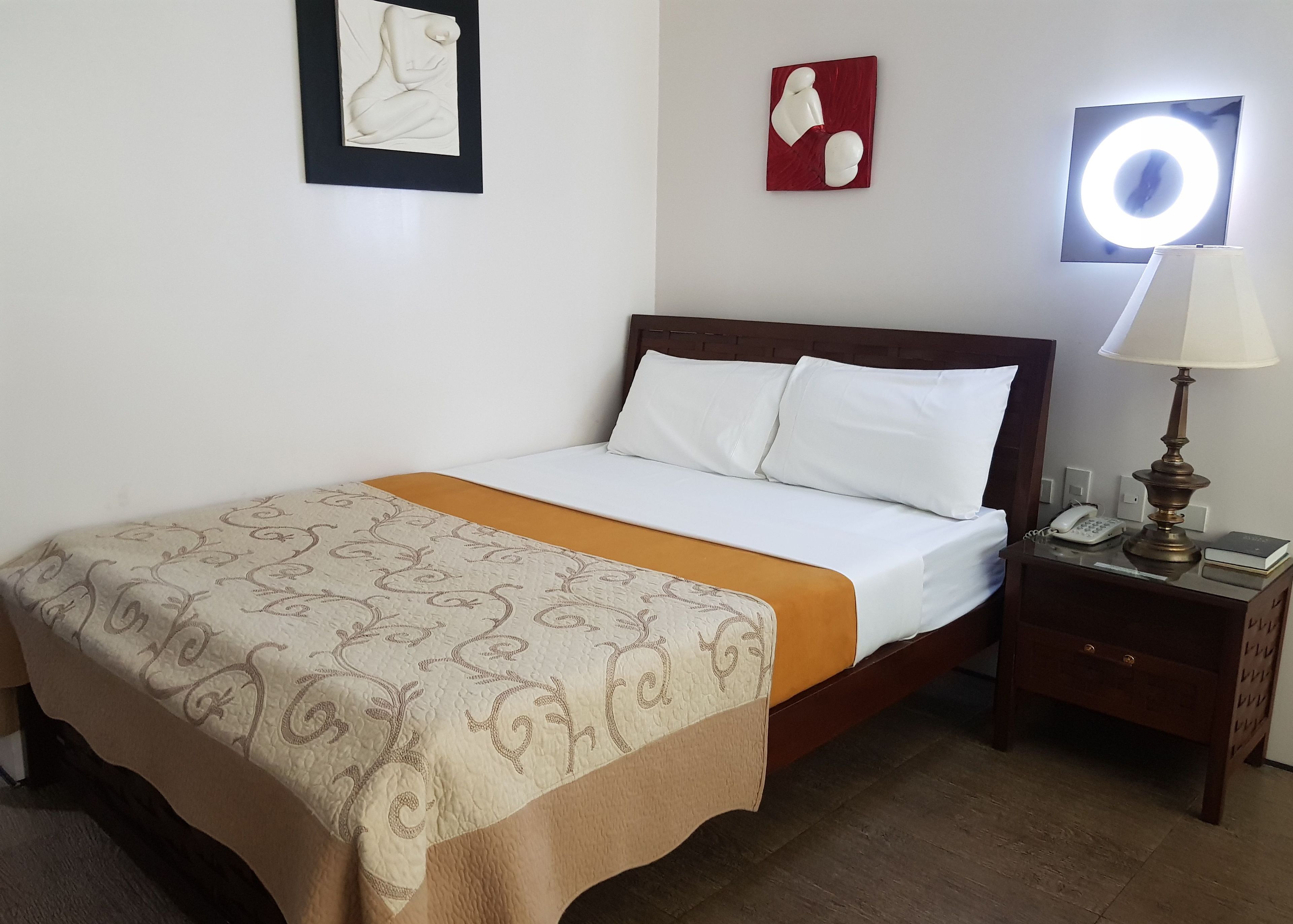 A neatly made bed with a patterned bedspread, a bedside lamp, a phone, and a small book on a wooden nightstand.
