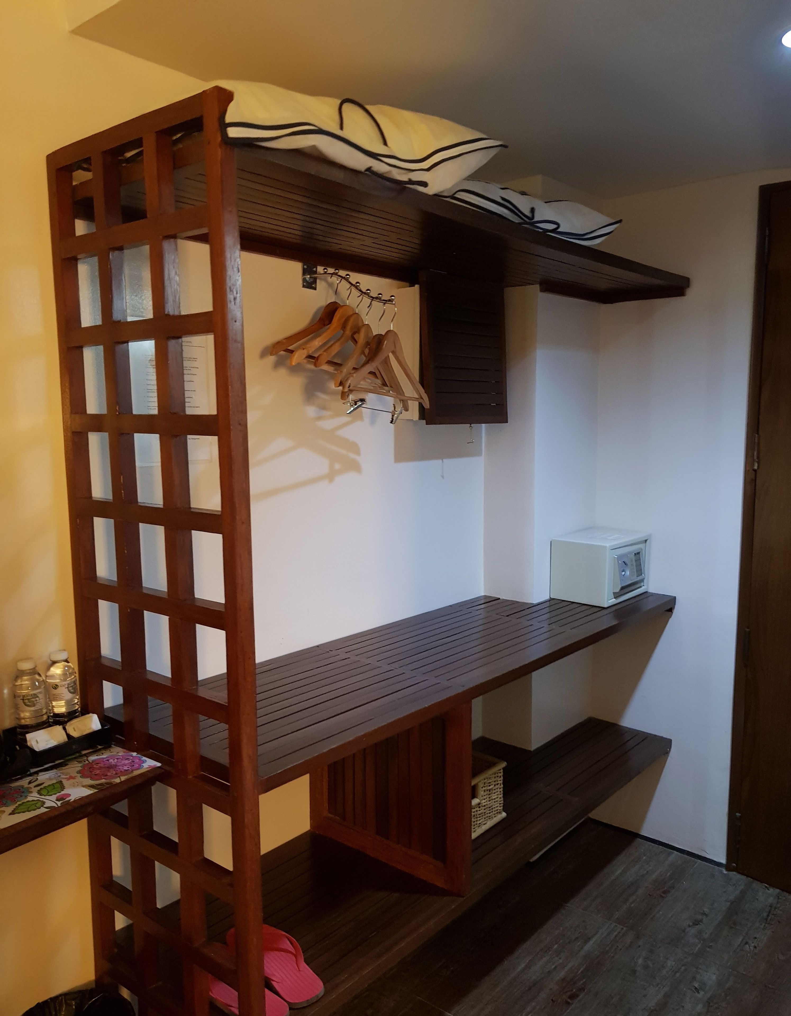 A small room with a wooden desk, a white microwave, and a wooden hanger rack with clothes.