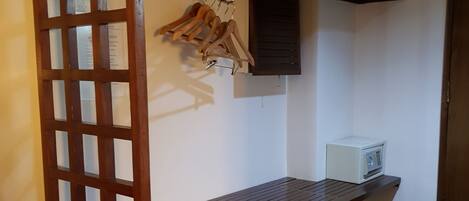 Quarto pequeno com escrivaninha de madeira, micro-ondas branco e um cabideiro de madeira com roupas.