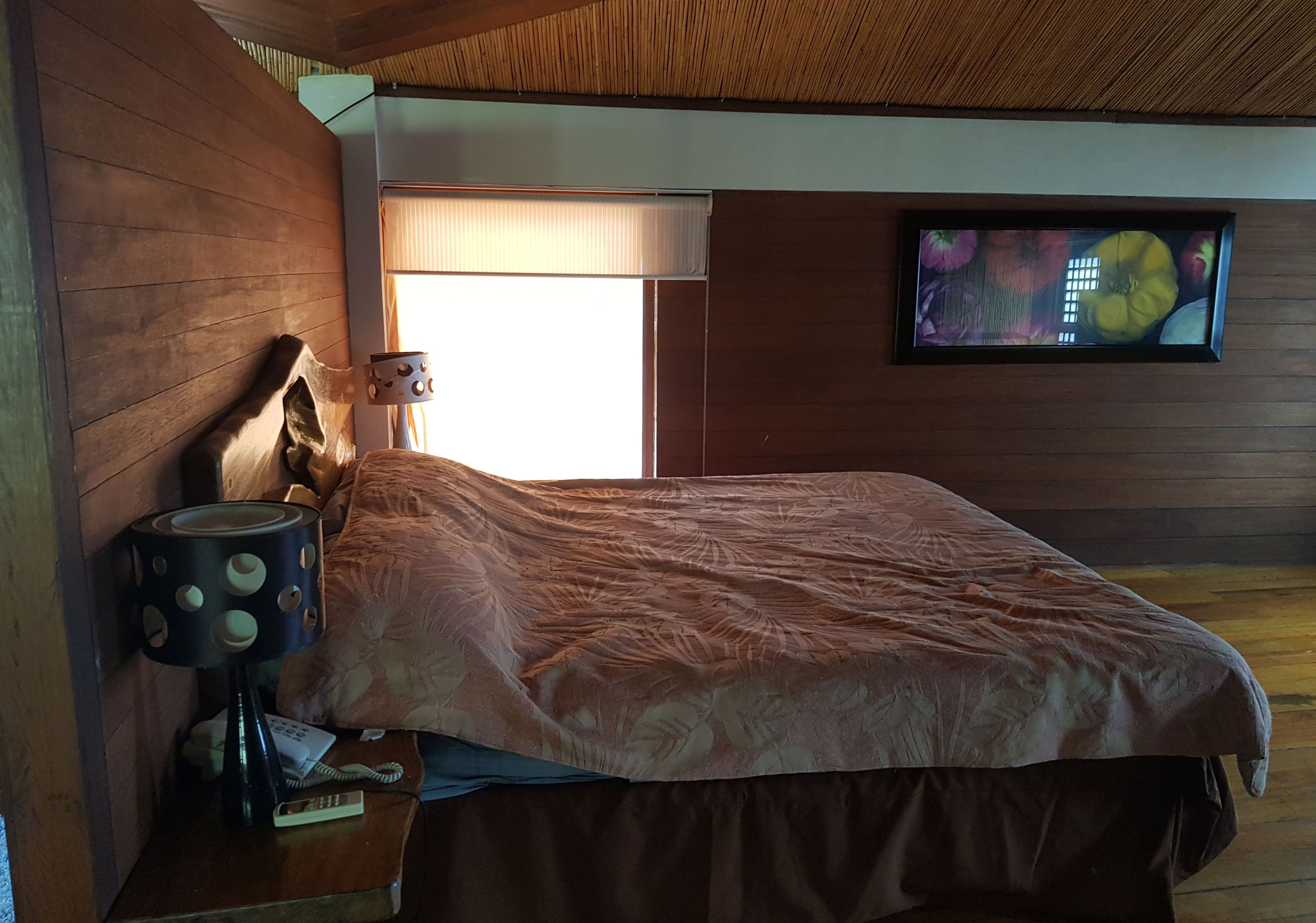 A bedroom with a bed, a nightstand with a lamp, a television on the wall, and a window with blinds.