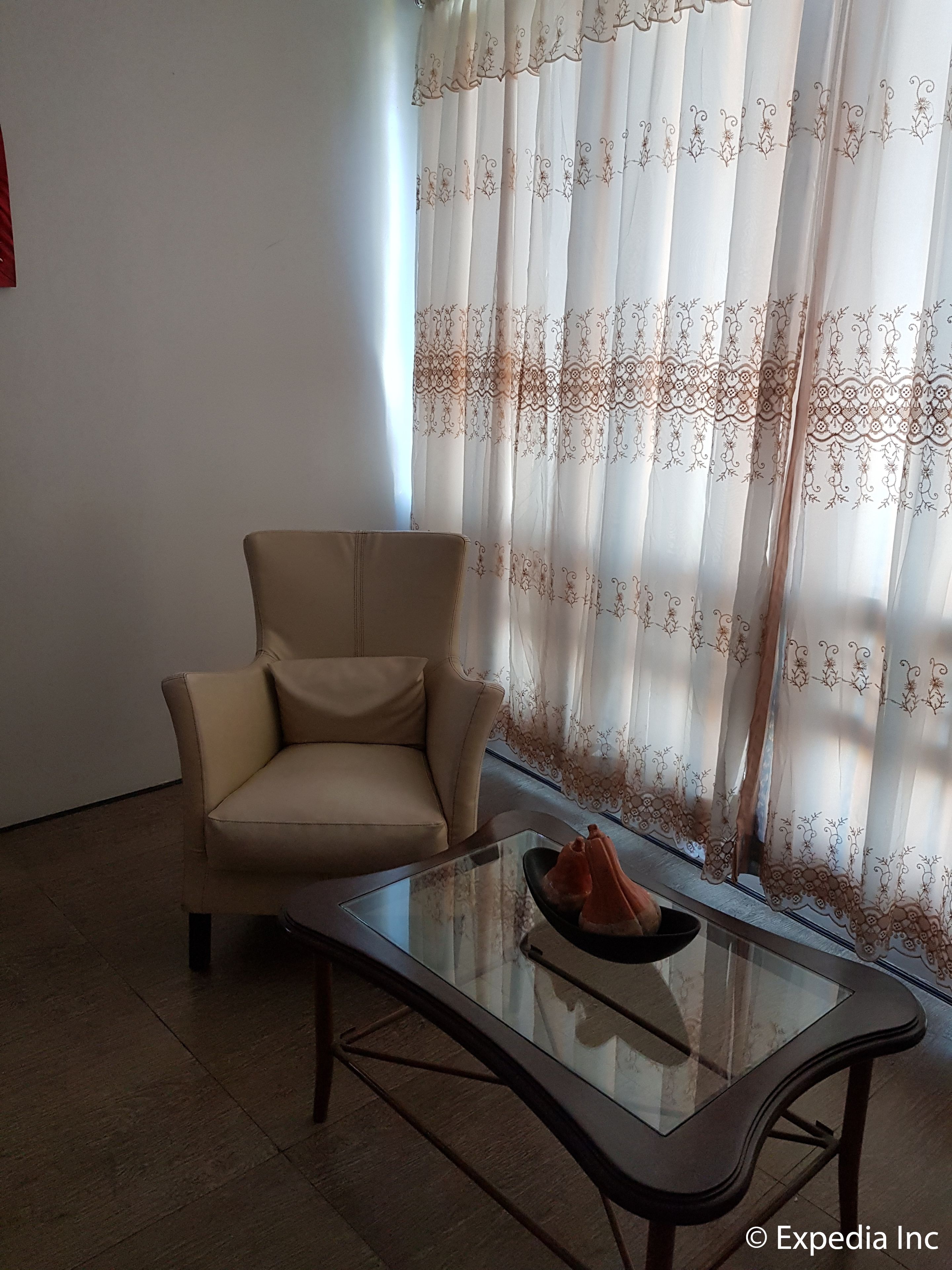 A beige armchair next to a glass-top table with a pair of slippers on it, in a room with a plain wall and a window with sheer curtains.