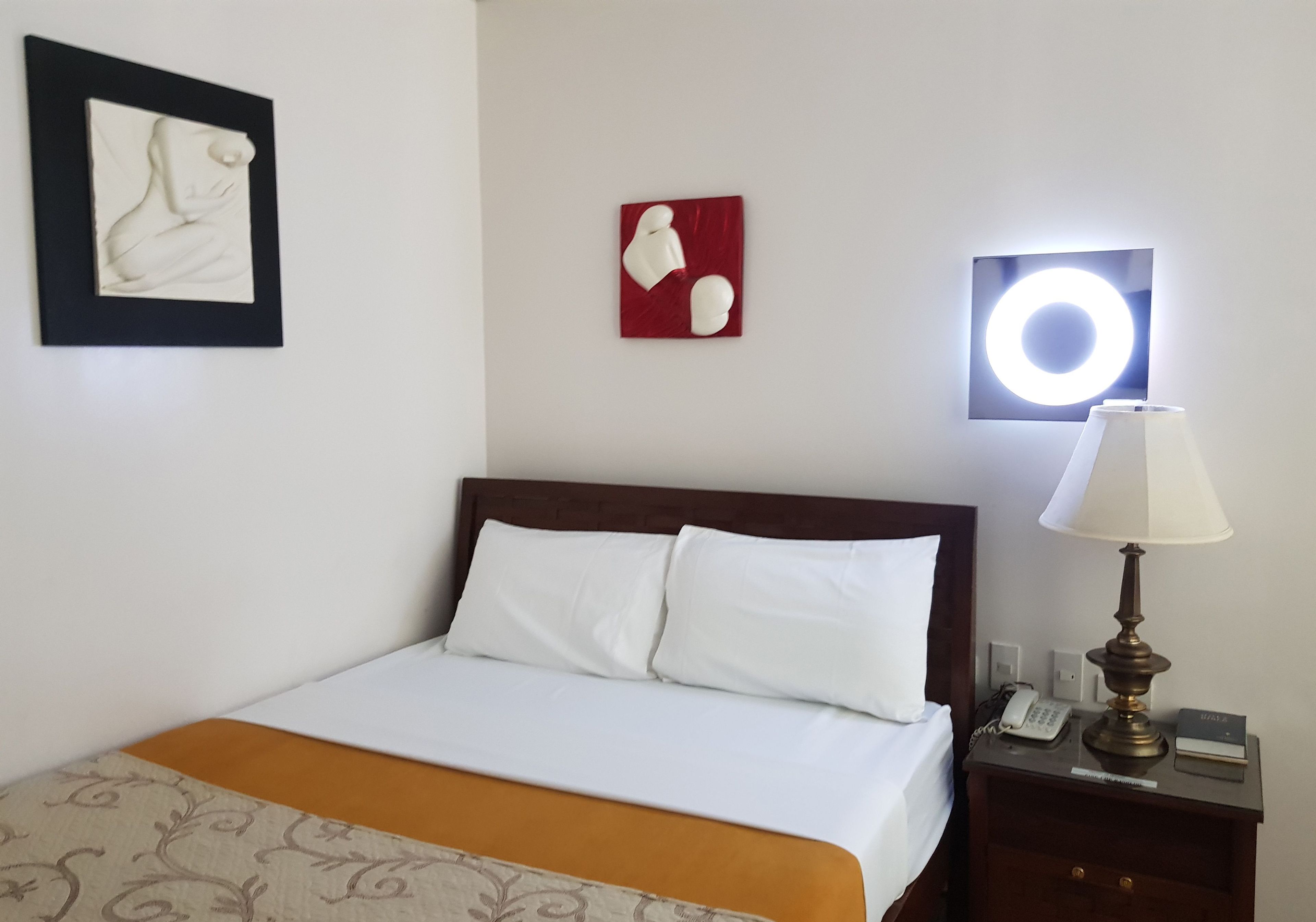 Una cama bien hecha con sábanas blancas y un plaid marrón, una lámpara de noche, un teléfono y una mesita con un libro y una placa de identificación.