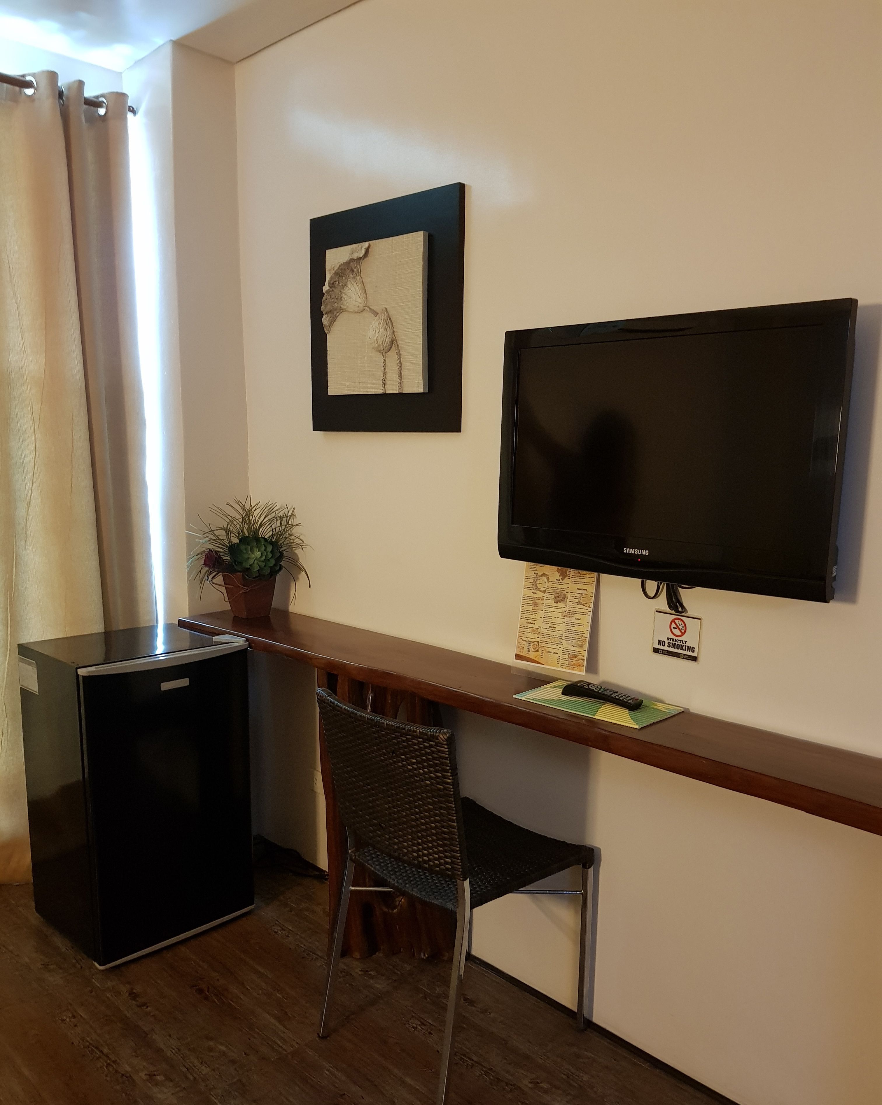 A compact room with a wooden desk, a black TV, a small fridge, a wicker chair, and a potted plant.