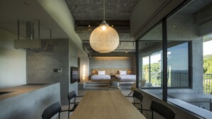 A modern hotel room with a large wooden table, black chairs, a hanging light fixture, a bed with white linens, and a balcony with a view of greenery.