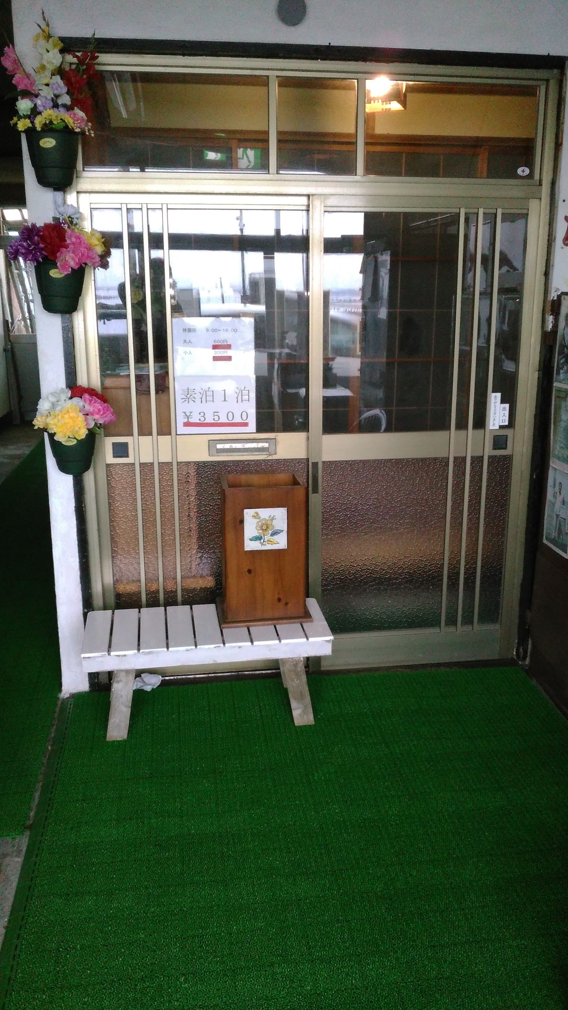 A hotel entrance with a green artificial turf, a wooden bench, and flower arrangements.
