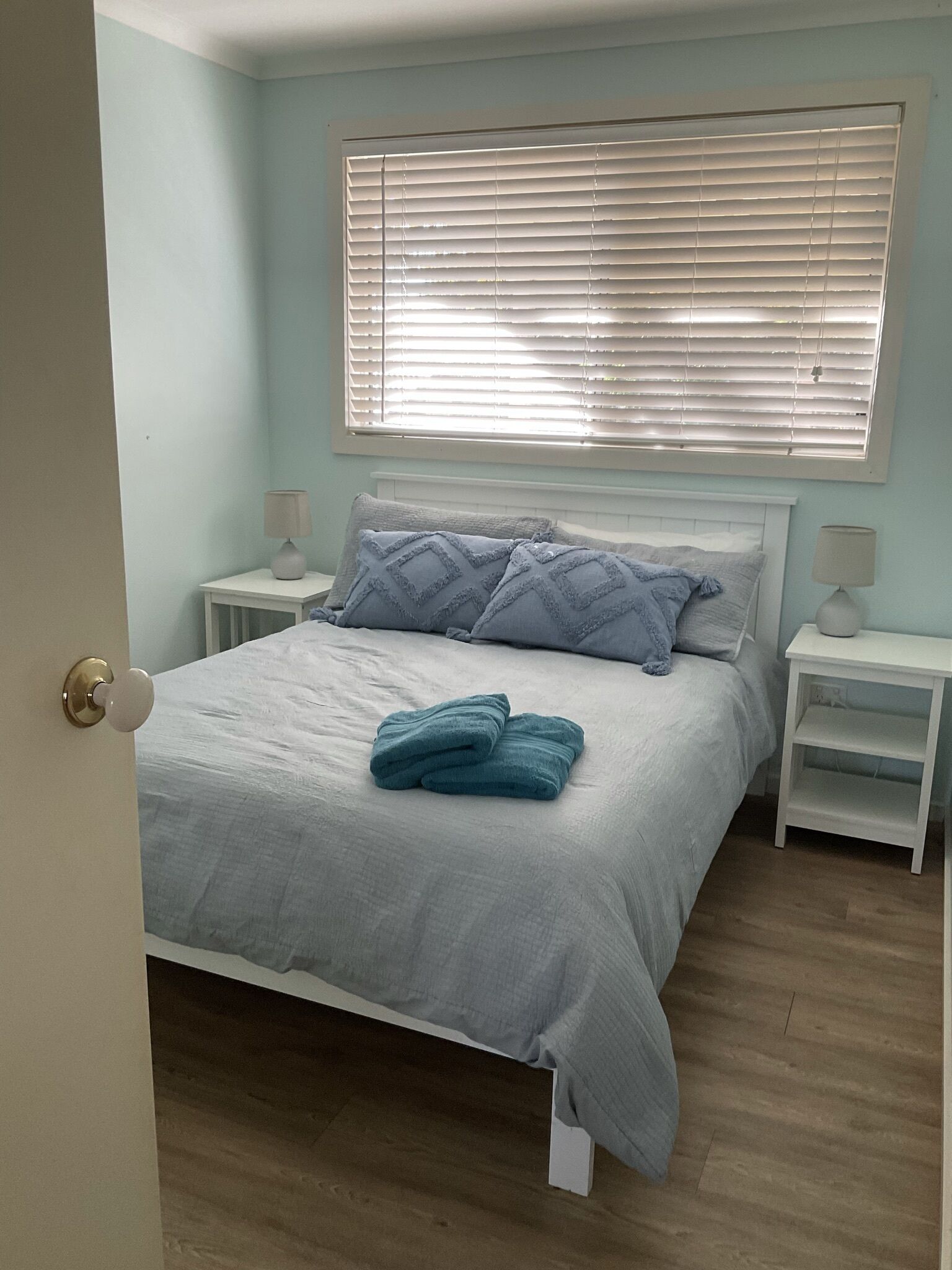 A bedroom with a bed, two bedside tables, a window with blinds, and a door.