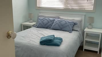 A bedroom with a bed, two bedside tables, a window with blinds, and a door.
