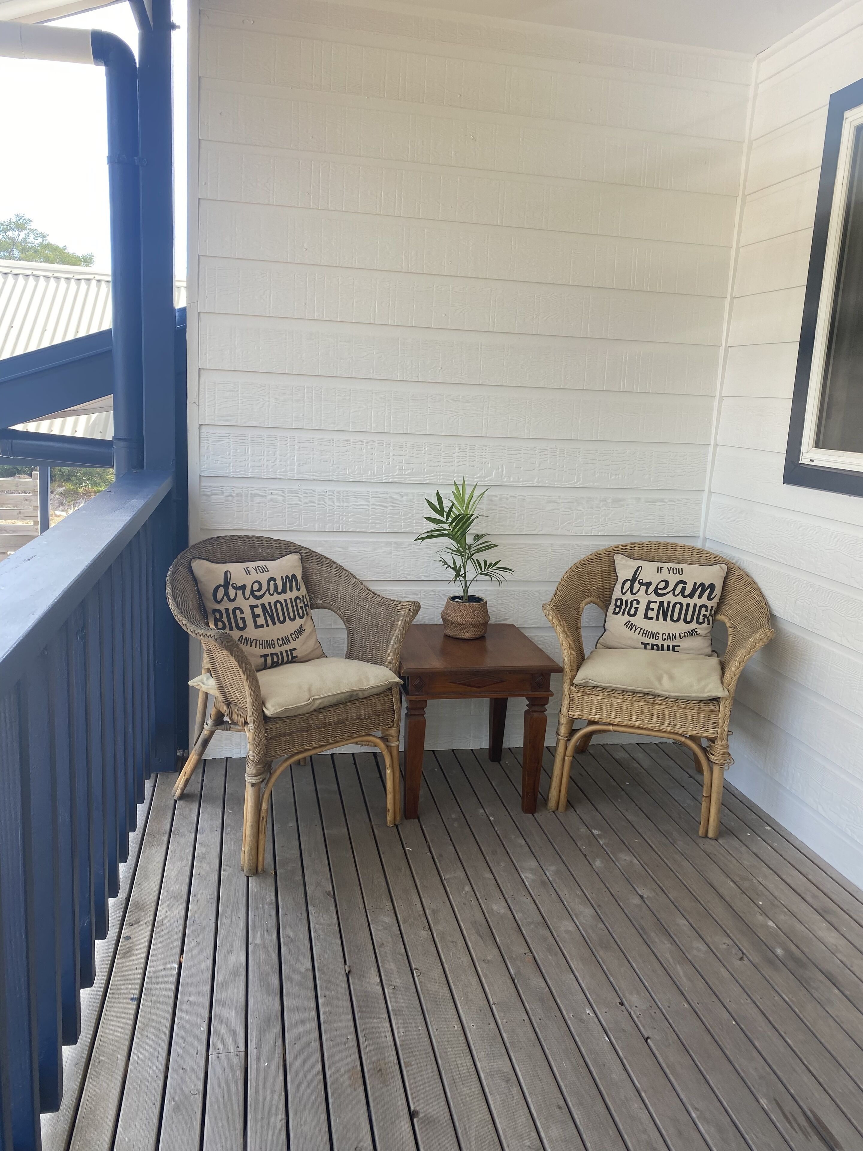 A wooden deck with two wicker chairs and a small table, each with a cushion and a pillow with the text "dream big enough anything can come true."