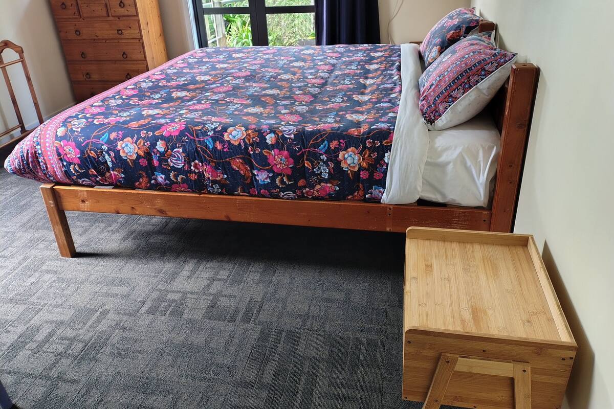 A wooden bed with a floral bedspread and matching pillows, a wooden nightstand, and a view of greenery outside.