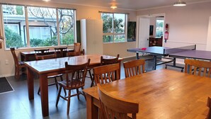 Una habitación amplia con mesa de ping pong, varias mesas y sillas de madera, ventanas grandes y una puerta con la señalización “EXIT”.