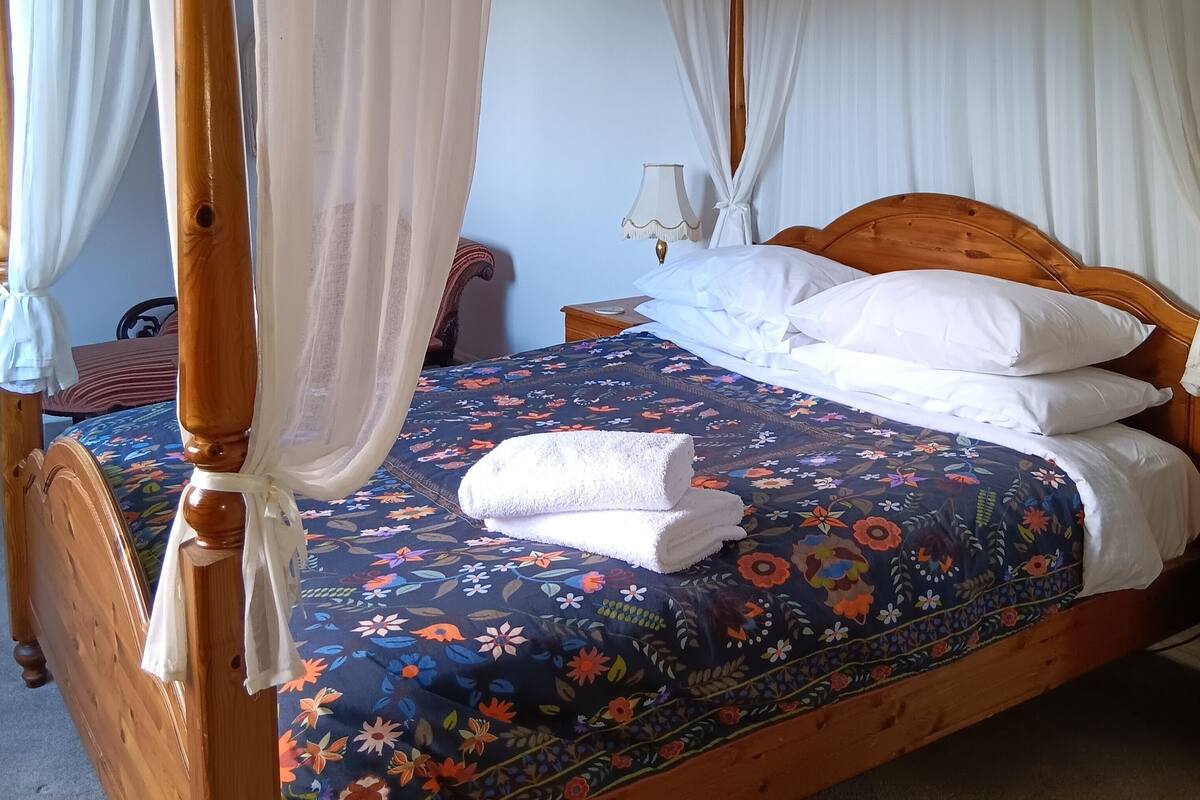 A four-poster bed with white curtains, a floral bedspread, and a neatly folded white towel.