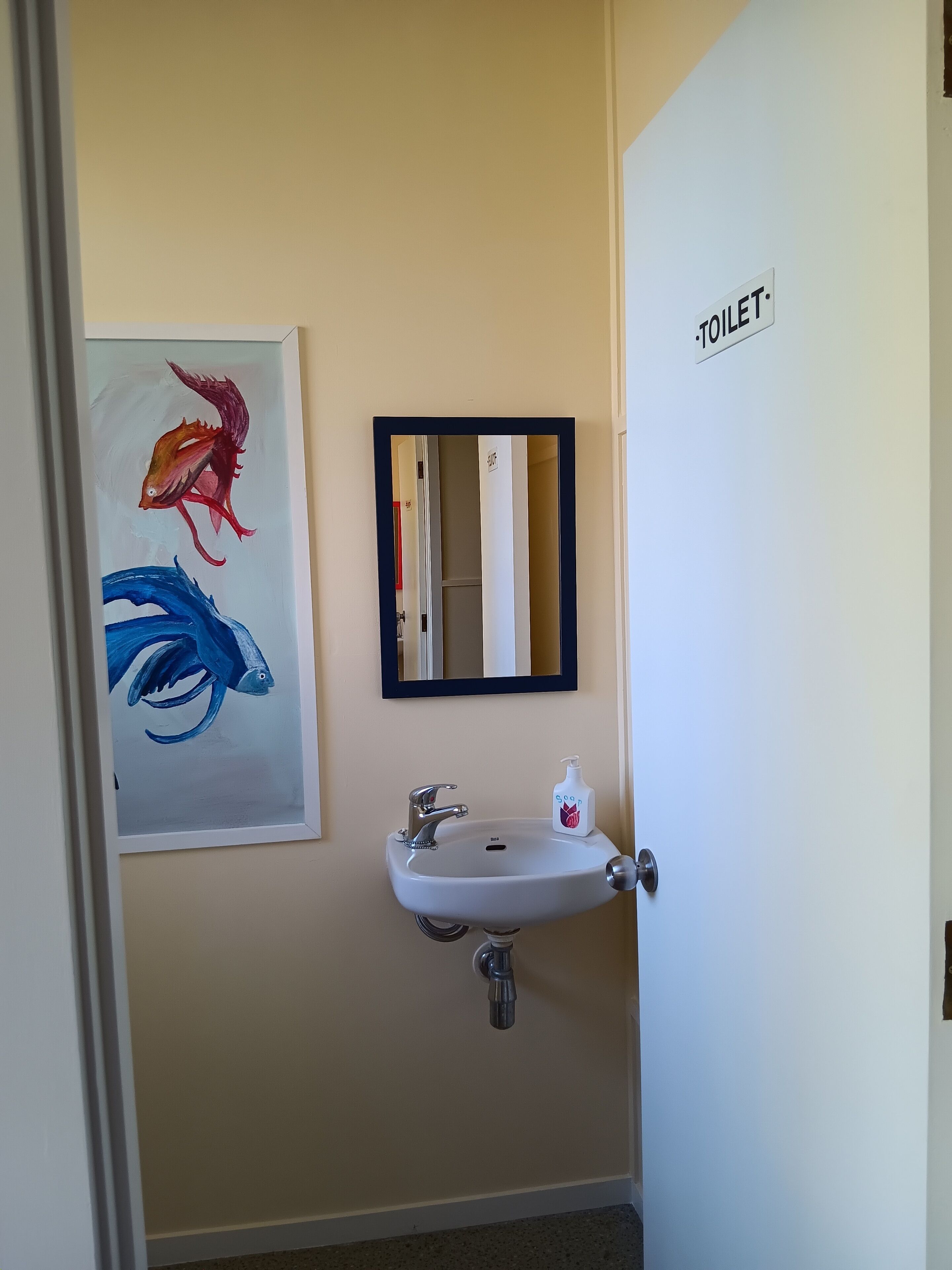 Un baño con lavamanos, espejo e inodoro. Hay un cuadro de un pez en la pared.