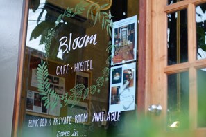 Janela com o texto "Bloom CAFE • HOSTEL" e "QUARTO EM CAMA BELICHE E QUARTO PRIVADO DISPONÍVEIS", indicando um estabelecimento tipo albergue.
