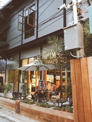 Un bâtiment moderne doté d’un espace café avec des places assises en plein air et d’un jardinier en bois où sont plantées des plantes.