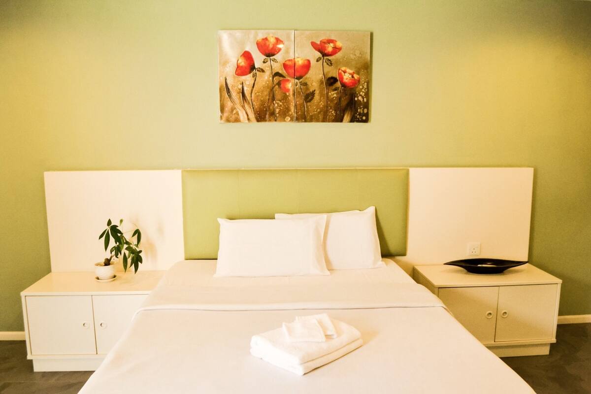 A hotel room with a bed, white bedding, a nightstand with a plant, a painting of flowers, and a bedside table with a bowl.