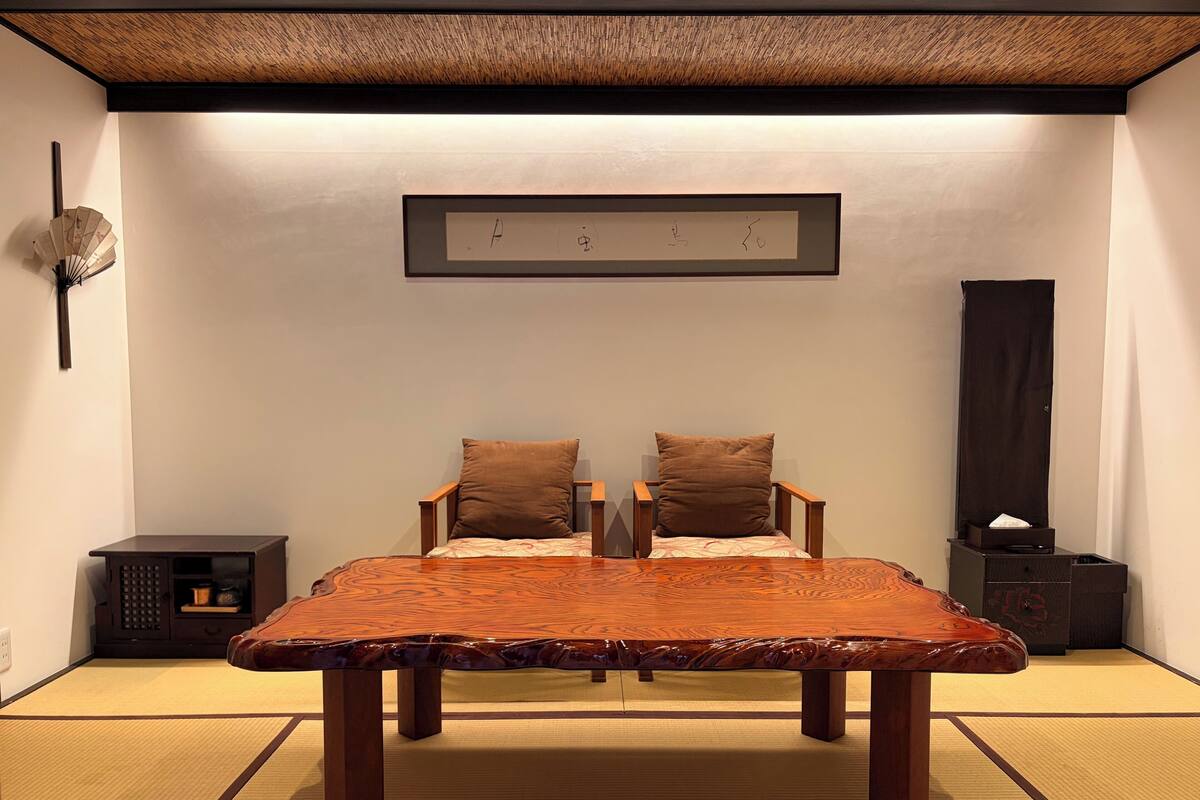 Une pièce de style japonais traditionnel, avec un sol recouvert de tatamis, une basse table en bois, deux chaises, un petit meuble et une œuvre d’art encadrée accrochée au mur.