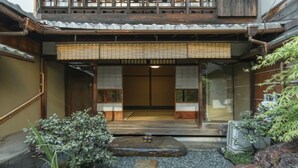 Ein traditionelles japanisches Gebäude mit Holzbeschichtung, einem Vorsprung aus Rattanmatten und einem kleinen Garten mit Steinen und Pflanzen.