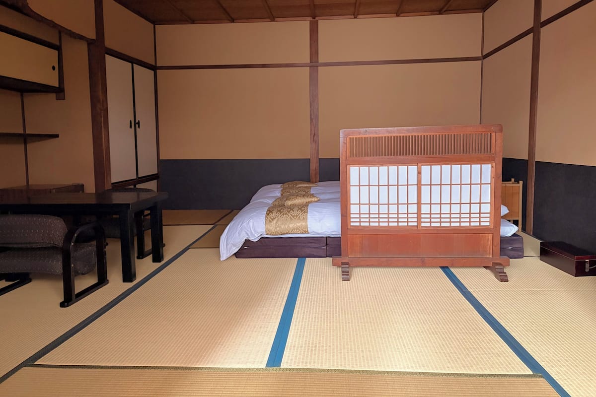 Une pièce de style japonais traditionnel, avec un sol recouvert de tatamis, un lit bas en bois muni d’un paravent coulissant, un bureau avec une chaise et une armoire.