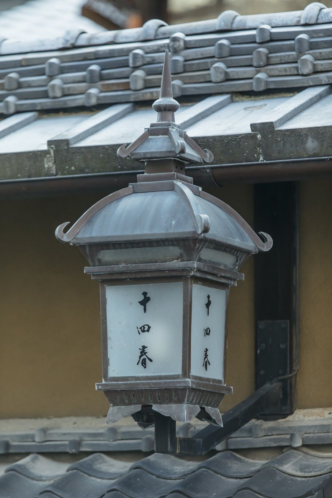 A traditional Japanese lantern with Chinese characters on its panels, mounted on a tiled roof.