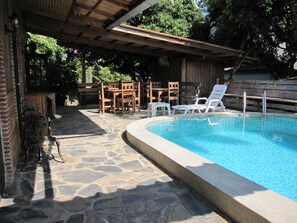 A stone-tiled pool area with a wooden pergola overhead, surrounded by wooden furniture and chairs. - XPAI Guesthouse (Pai)