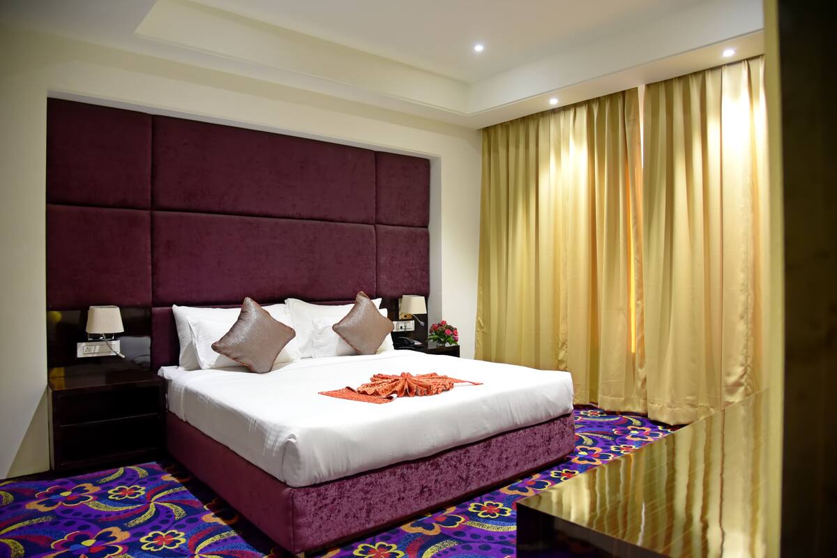 A hotel room with a large bed, a headboard, a nightstand, a lamp, and a window with curtains.