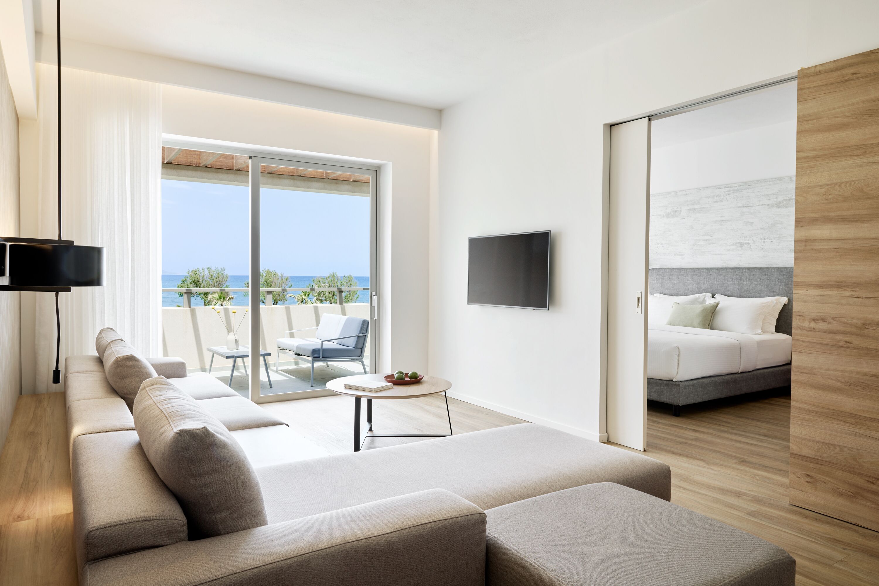 Ein modernes Wohnzimmer mit einer Couch, einem kleinen Tisch und einem Fernseher. Eine Schiebetür führt zu einem Balkon mit Blick aufs Meer.